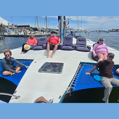 Six people sit and lounge on the deck of a catamaran docked at a marina, with water and buildings visible in the background under a partly cloudy sky.