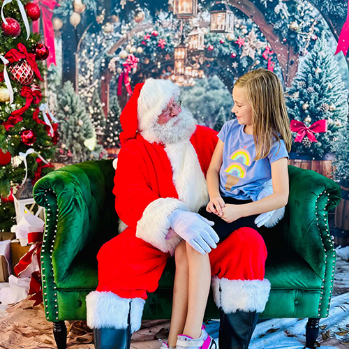 A girl sits on Santa Claus&rsquo;s lap on a green couch, surrounded by Christmas decorations and a decorated tree in the background.