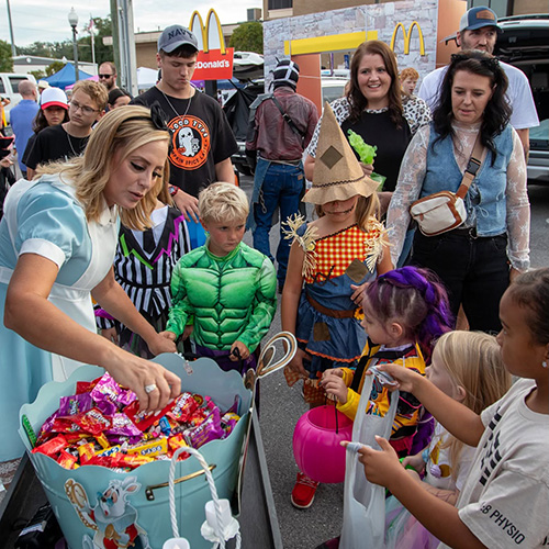 A woman hands out candy to children in costumes at an outdoor event, with adults and kids gathered around and a McDonald&rsquo;s restaurant visible in the background.