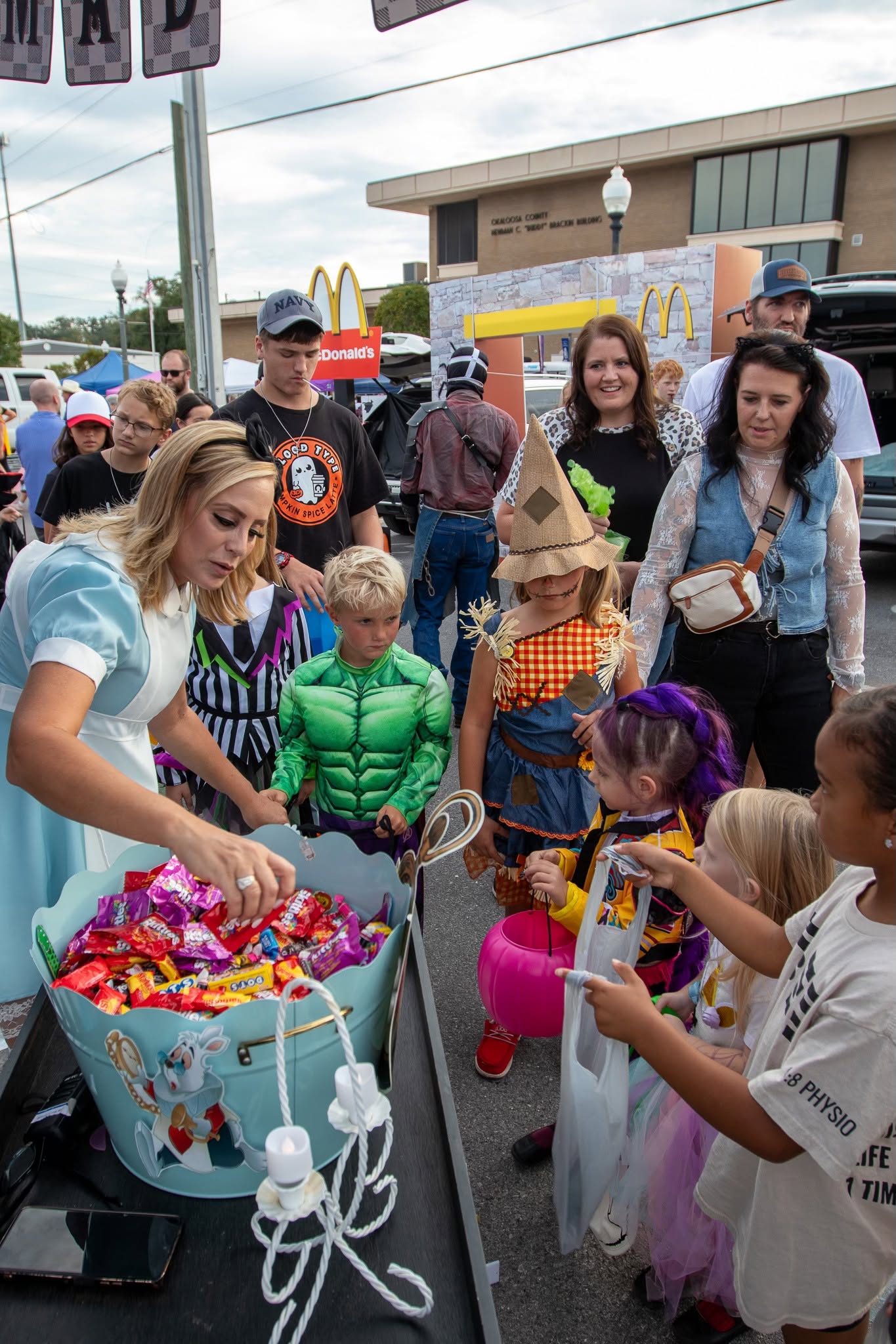 A woman hands out candy from a bucket to children in costumes at an outdoor event, while adults and other kids watch.