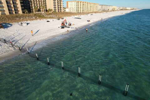 Aerial view of a sandy beach with a few people, beach tents, and posts in the shallow turquoise water near shoreline, with hotels in the background.