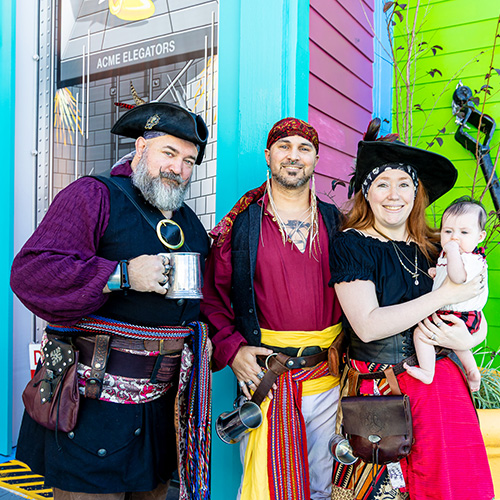 Four people dressed in pirate costumes, including one holding a baby, stand smiling in front of a colorful building.