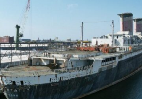 A large, rusted ship sits docked at a port under a clear sky, showing signs of age and disrepair.