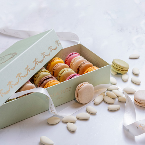 A light green box filled with assorted macarons, surrounded by white almonds and a ribbon.