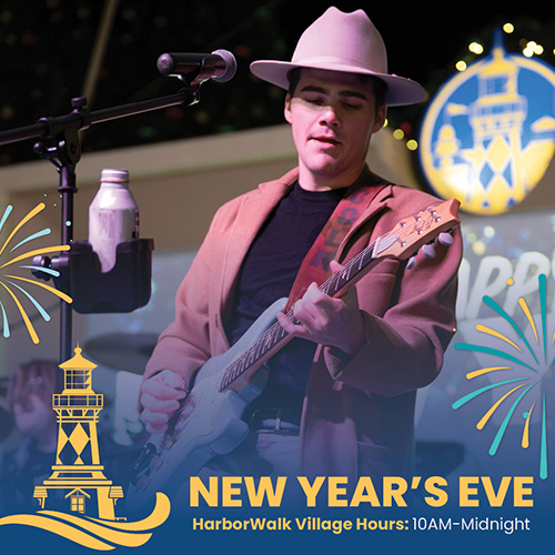 A man in a hat plays an electric guitar on stage, with overlaid text reading &ldquo;New Year&rsquo;s Eve, HarborWalk Village Hours: 10AM-Midnight.&rdquo;
