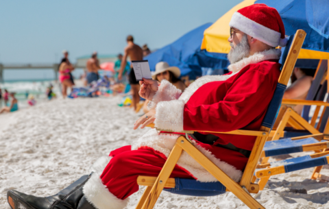 Santa Claus in a red suit sitting in a beach chair on the sand, reading a small card.