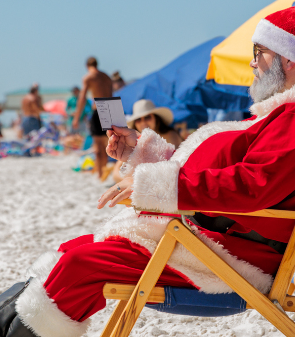 Santa Claus in a red suit sitting in a beach chair on the sand, reading a small card.